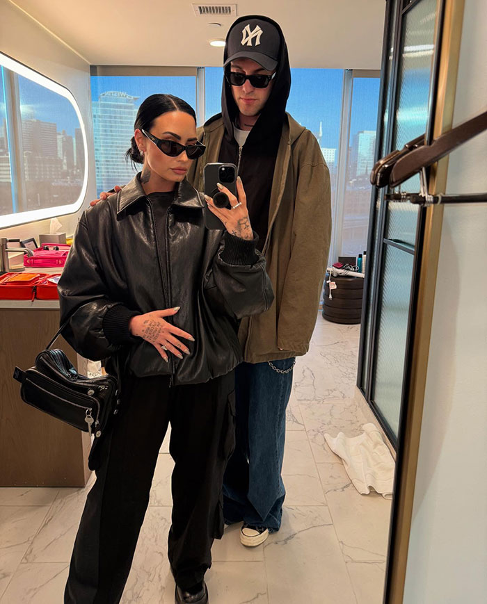 Two people in a modern room, one wearing a leather jacket, another a hat, both with sunglasses, reflecting "skinny" frame.