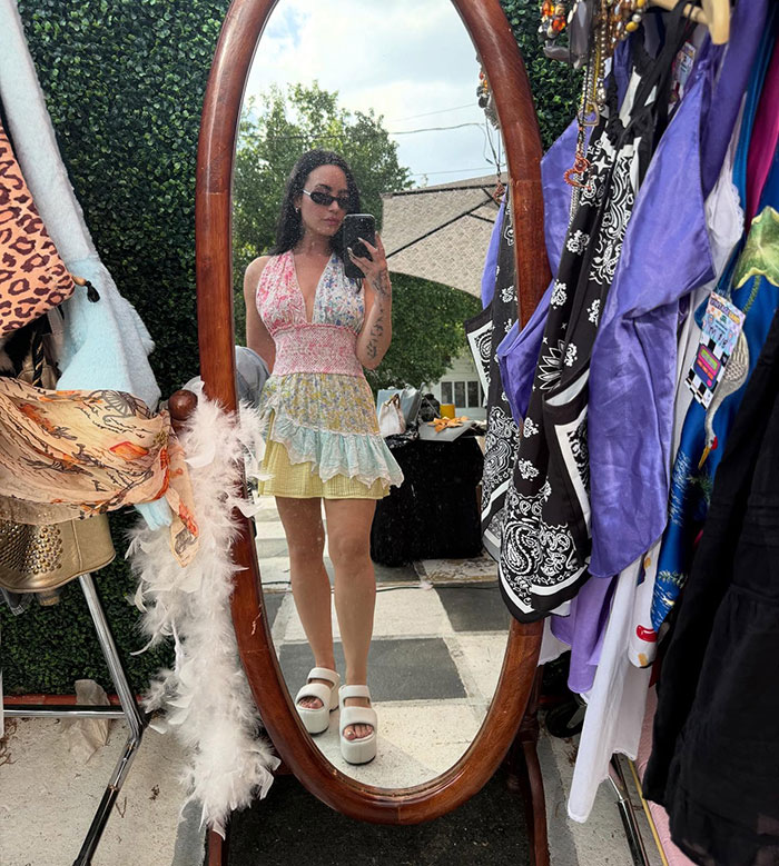 Person in summer dress and sunglasses taking a mirror selfie, surrounded by colorful clothing.