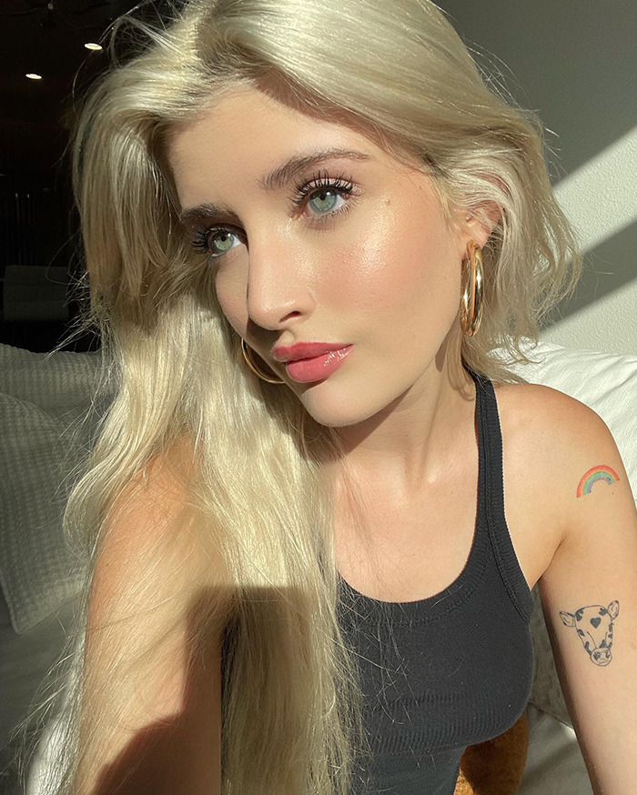 Young woman with long blonde hair, wearing a black tank top, indoors with sunlight on her face, discussing cosmetic surgeries.