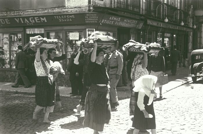 Women carrying baskets on their heads walking a cobblestone street showing life in Europe 100 years ago.