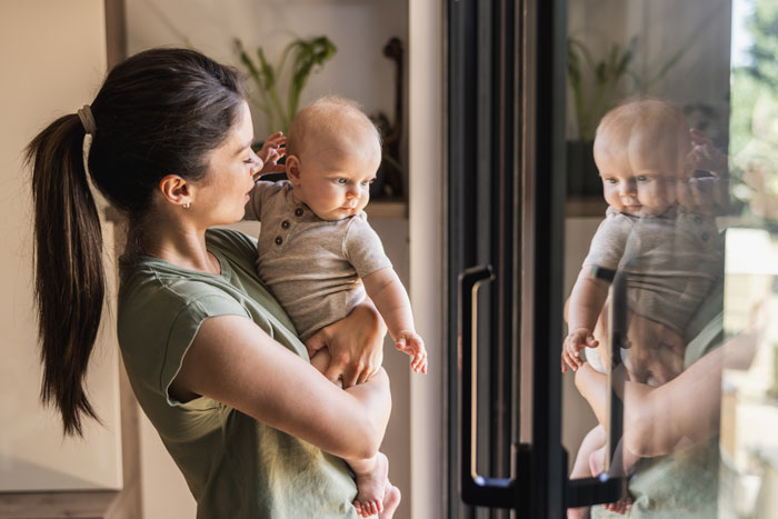 Woman holding baby near window, baby looking outside, reflecting family dynamics.