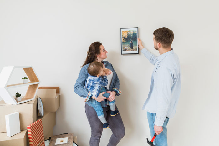 A woman holding a baby watches a man hanging a photo on the wall, boxes in the background suggest a home setup.
