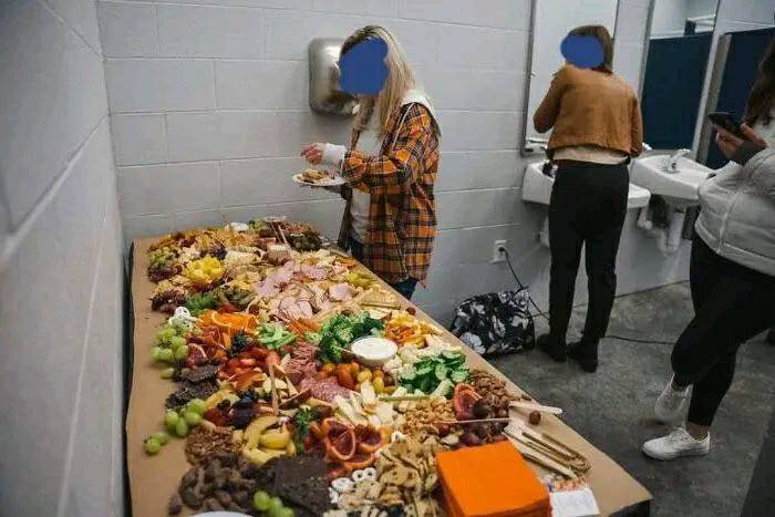 Potluck dish spread in a restroom setting, featuring assorted meats, cheeses, fruits, and snacks on a table.