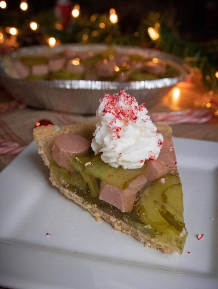 Unusual potluck dish with pie topped with hot dogs, pickles, and whipped cream, surrounded by holiday lights.