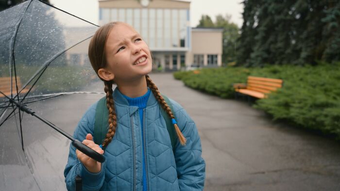 Child with braids, holding a clear umbrella, looking up and smiling in a park, proving to be a true comedian.