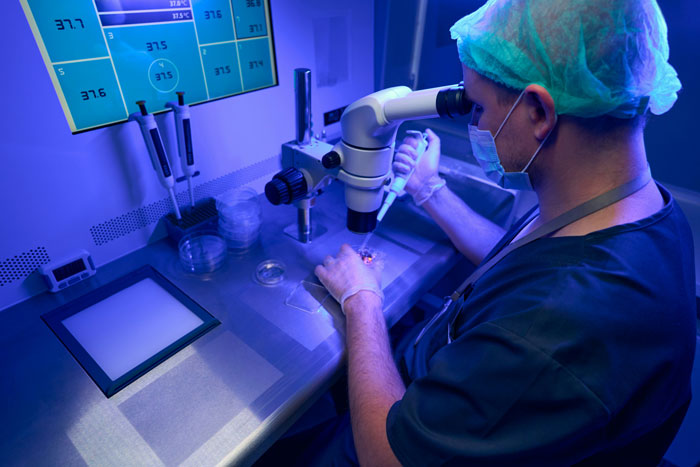 Scientist in lab using microscope for fertility research, wearing blue scrubs and hairnet.