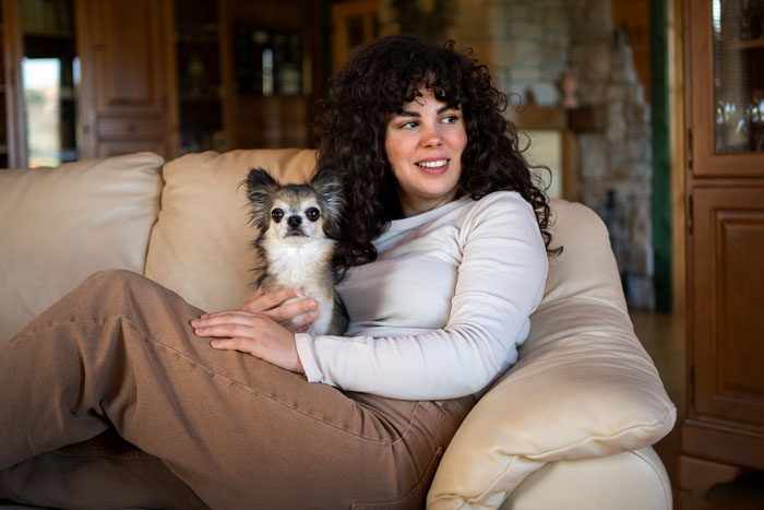 A woman with curly hair lovingly holds a small dog on a beige sofa, highlighting emotional support and hope.