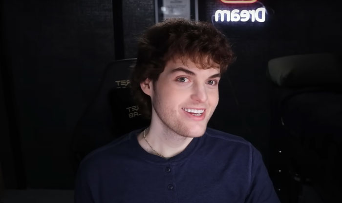 Youtuber Dream with curly hair smiling in a dark room with a glowing Dream sign, representing Minecraft star&rsquo;s face reveal moment.