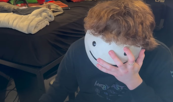 Youtuber Dream with curly hair covering face with hand, wearing a white smiley mask, related to Dream face reveal and Minecraft star fans.