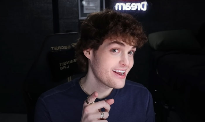 Youtuber Dream smiling and pointing, sitting in a gaming chair with Dream neon sign in the dark room.