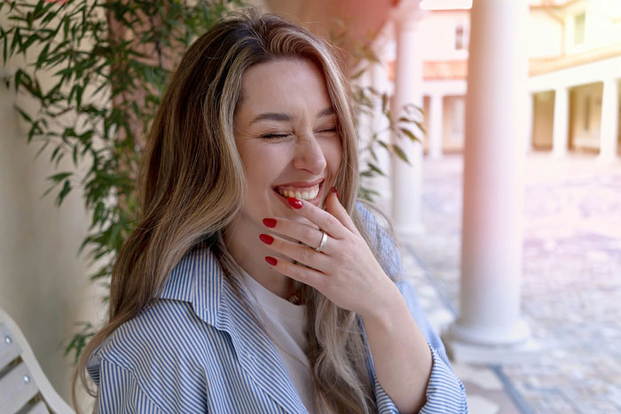Smiling woman with red nails wearing a ring, enjoying a sunny day near a columned walkway outdoors.