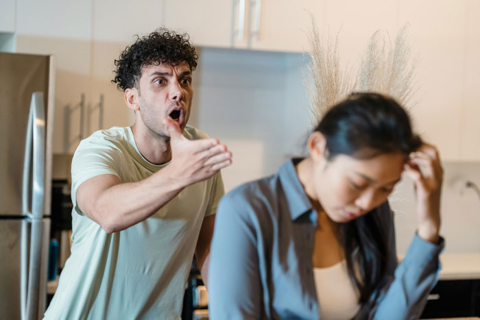 Man upset in kitchen, gesturing with hand, while woman looks down.