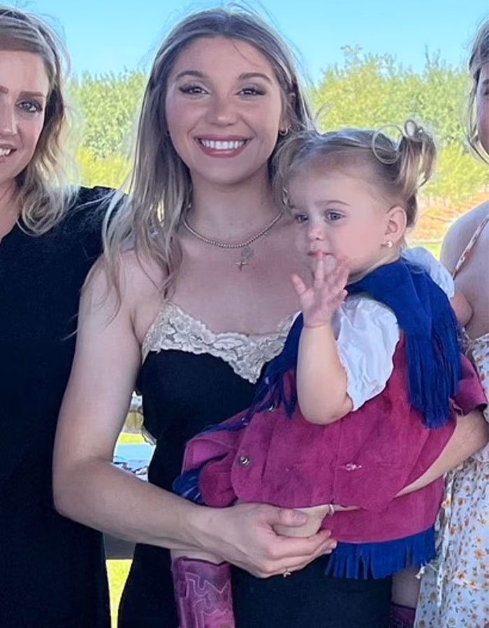A mother holding her young daughter, both smiling outdoors.