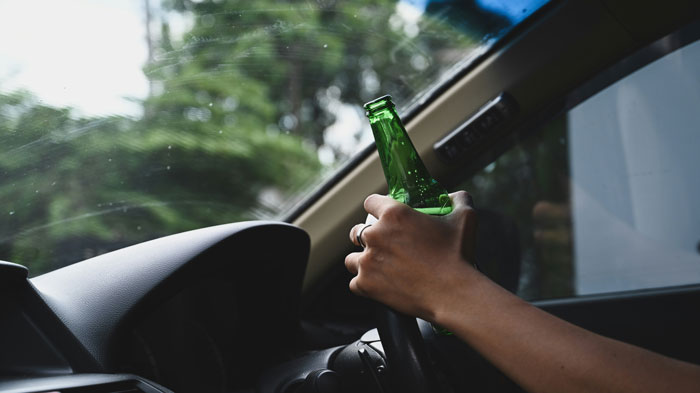 A hand holding a green bottle while steering a car, highlighting drunk driving.