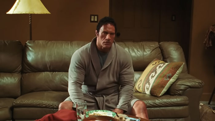Dwayne Johnson showing impressive transformation in a robe, sitting on a couch preparing for new biopic role.