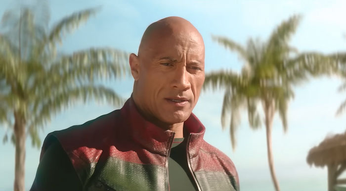 Dwayne Johnson showing impressive transformation in a red and green jacket, standing outdoors with palm trees in the background.