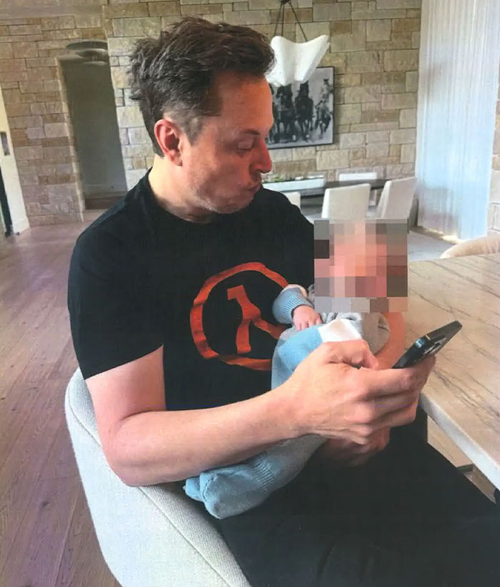 Man sitting in a chair, looking at his phone, holding a baby with a blurred face.