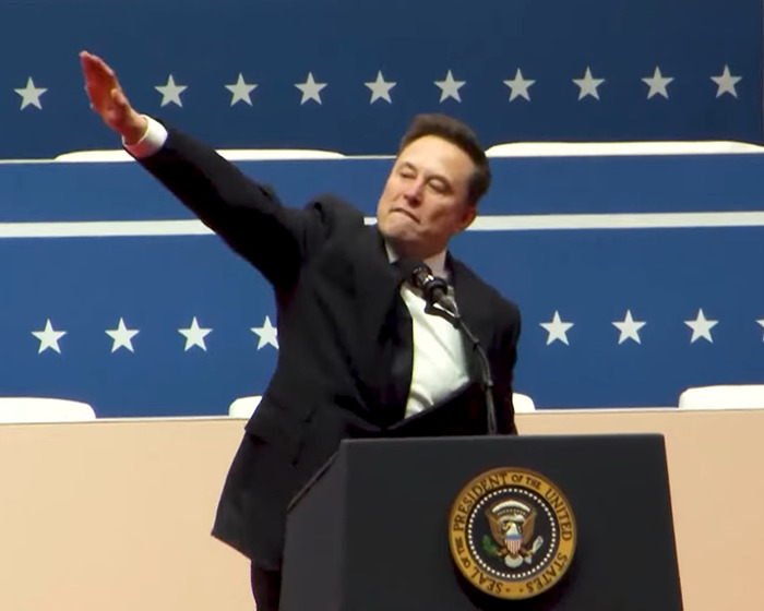 A person in a suit gesturing at a podium, with blue star-patterned backdrop behind them.