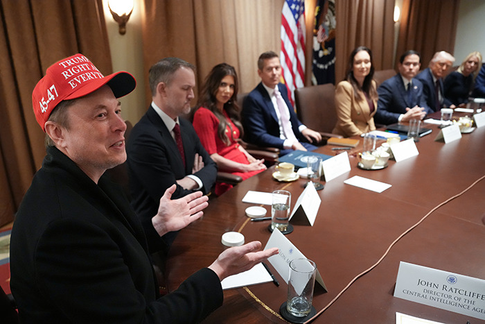 Person in a red hat discussing at a conference table with several attendees, focusing on DOGE topic.