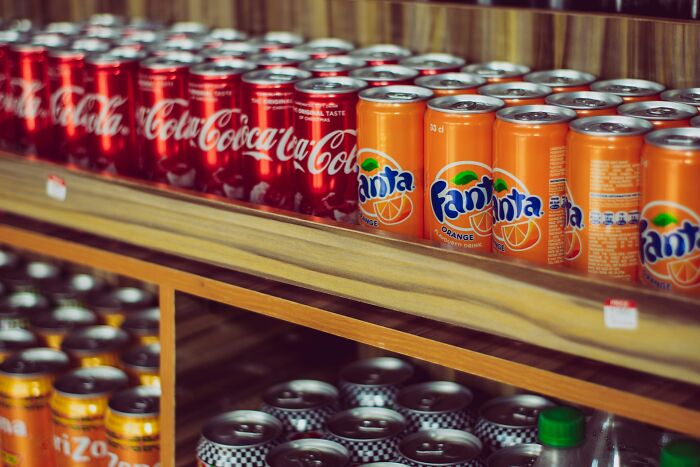 Rows of Coca-Cola and Fanta soda cans on wooden shelves in a restaurant food setting with beverage products displayed.