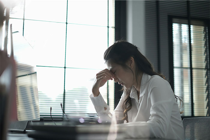 Employee looking stressed at her desk, holding her forehead in an office environment.