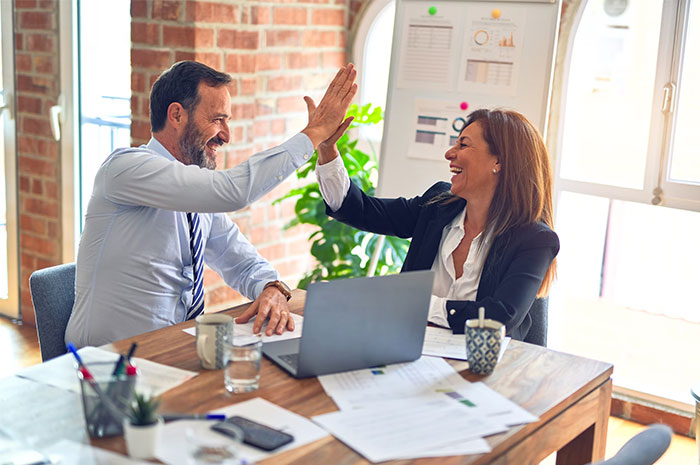 Two colleagues high-fiving at a desk with a laptop and paperwork, illustrating workplace dynamics and hidden insights.