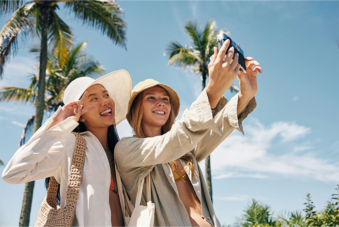 Two women taking a selfie under palm trees, capturing a fun moment in tropical setting.