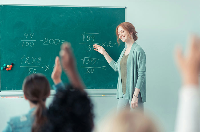 Teacher smiling while explaining math on a chalkboard in a classroom setting, with students raising hands.