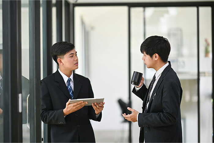 Two employees in discussion, one holding a tablet, the other a mug, in an office setting.