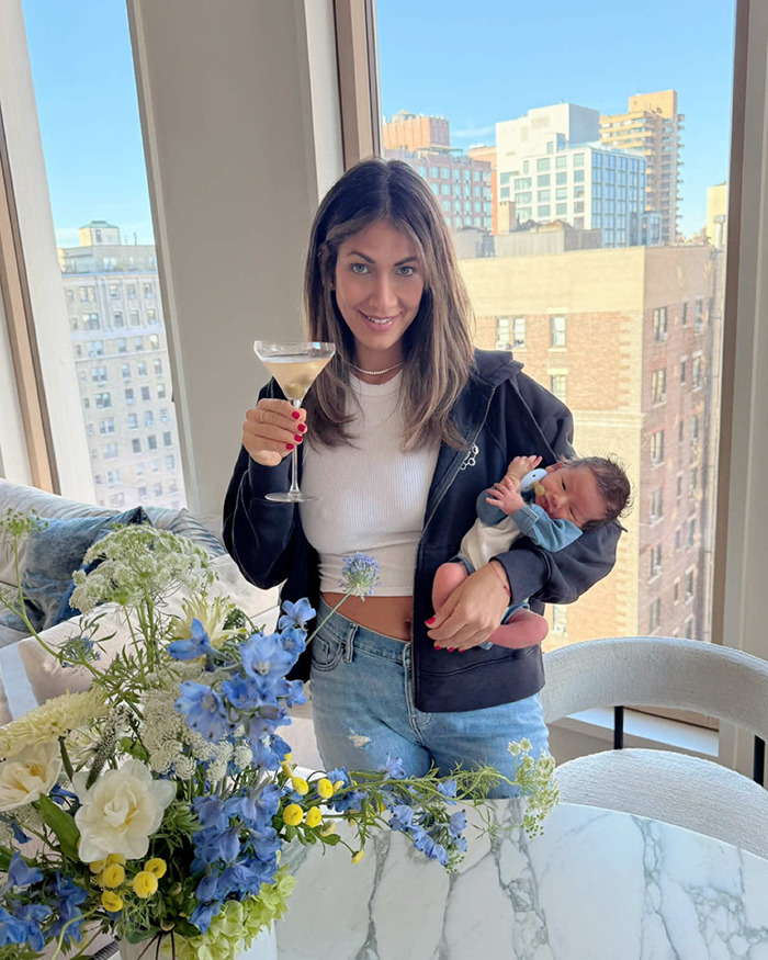 Woman holding a baby and martini near a window, surrounded by flowers, sparking safety concerns.