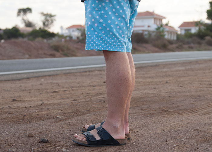 Man wearing blue polka dot shorts and sandals, standing beside a rural road, possibly an American traveler.