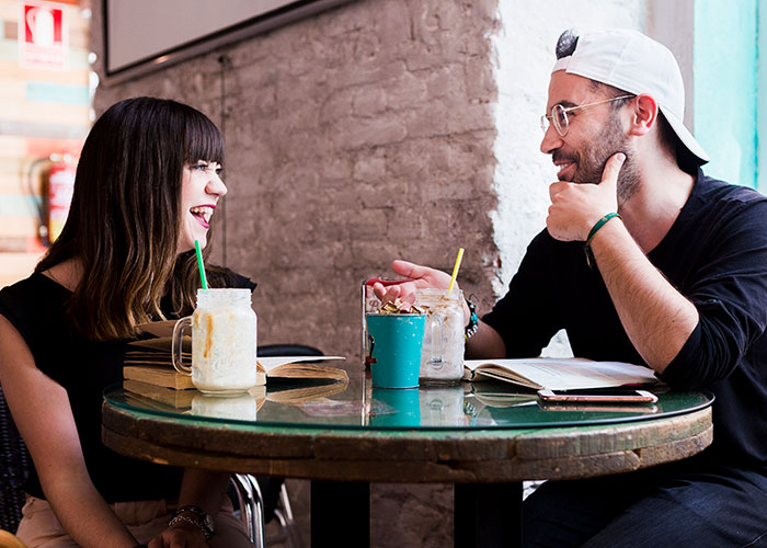 Two friends laughing at a cafe table, possibly discussing American travel experiences in Europe.