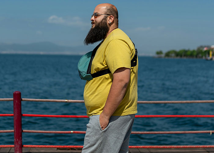 Man with beard in yellow shirt and f***y pack, standing by a lakeside, illustrating American traveler traits in Europe.