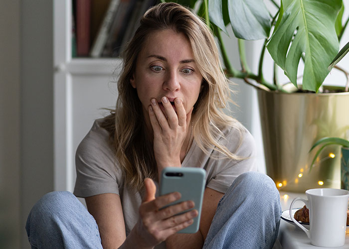 Woman looking surprised at phone, sitting with plants and coffee, revealing you're American.