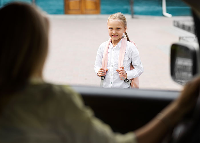 A young girl with a backpack smiles while approaching a car, viewed from inside, hinting at American cultural traits.