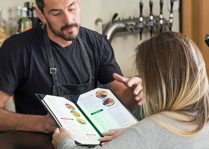 A server and customer discussing a menu at a European restaurant, highlighting cultural travel differences.