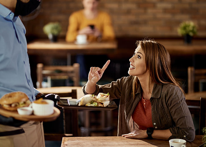 Woman pointing while talking to a waiter in a café, highlighting American tourist behavior.