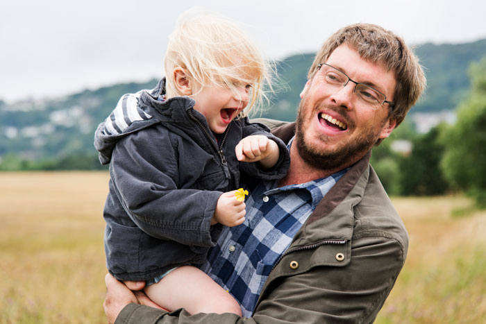 A man happily holds a child in an open field, both smiling and enjoying the moment.