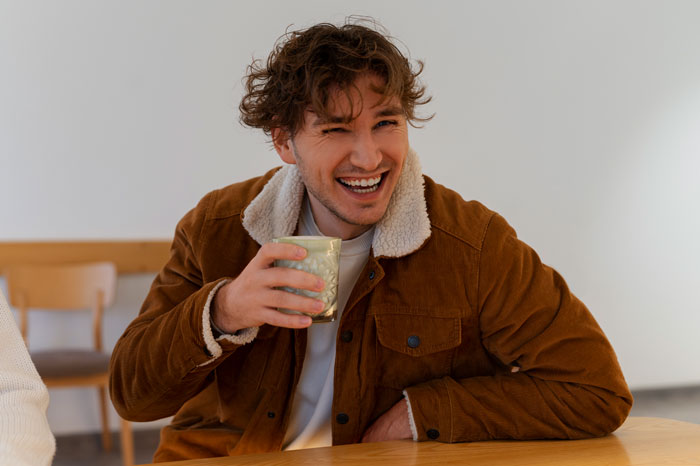 Man laughing with a drink at a table, feeling cheerful despite being dumped before Valentine's.