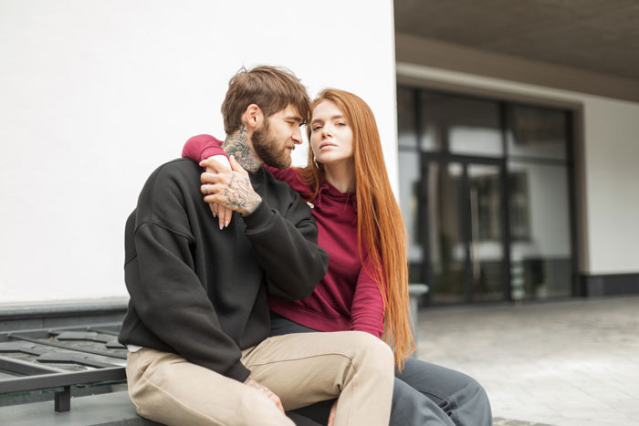 Man with tattoos and woman sitting closely on a bench, sharing an intense moment before Valentine's.