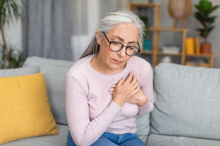 Elderly woman sitting on a couch, holding her chest and looking concerned.