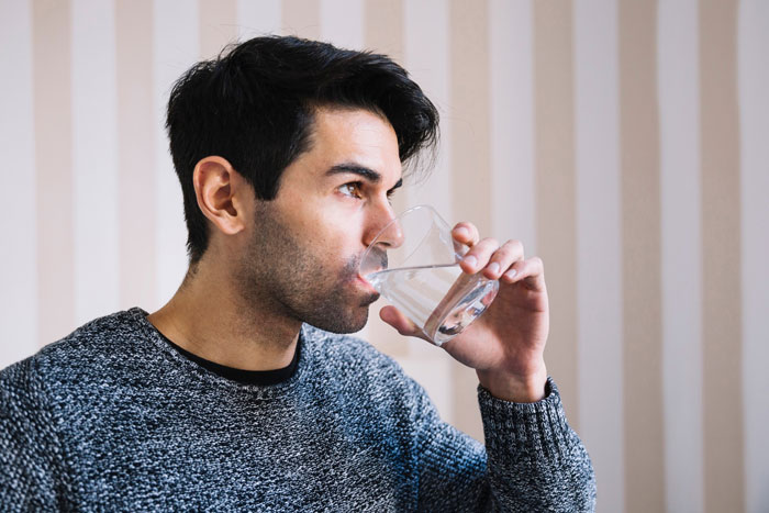 Man drinking sparkling water from a glass with a contemplative expression, near striped wallpaper.
