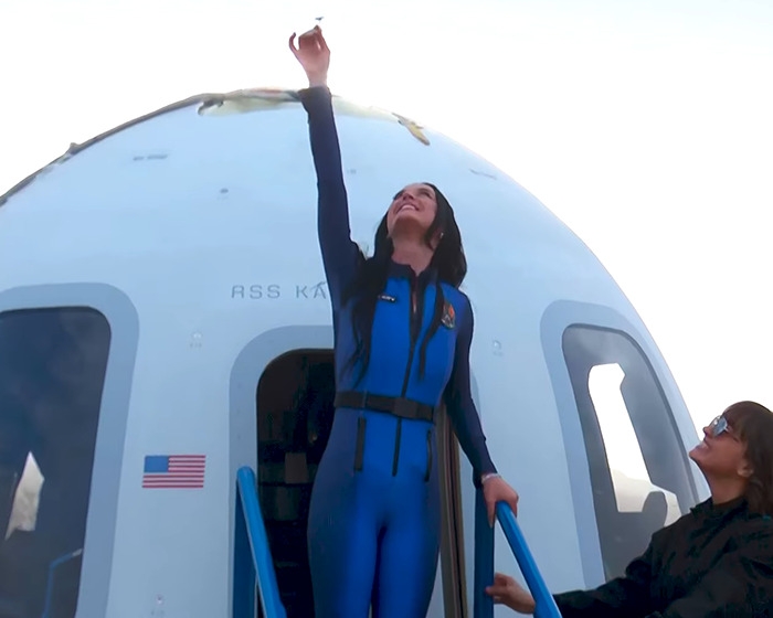 Individual in a blue spacesuit standing on steps of a spacecraft, reaching upwards, smiling. Individual in a blue spacesuit standing on steps of a spacecraft, reaching upwards, smiling.