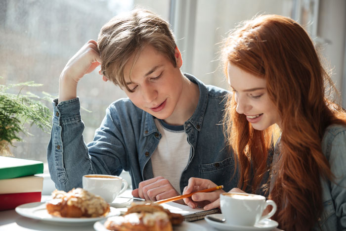 Teen couple smiling at a table with coffee, disregarding dad's ban due to grades slipping.
