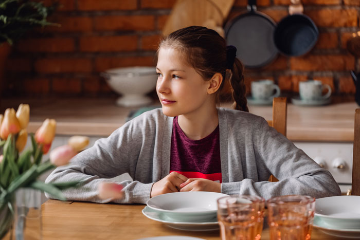 A young girl, possibly the daughter, sitting at a table set with plates and glasses, looking towards the side.