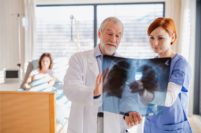 Doctor and nurse reviewing lung x-ray while woman lies in hospital bed dealing with physical consequences of accident Doctor and nurse reviewing lung x-ray while woman lies in hospital bed dealing with physical consequences of accident