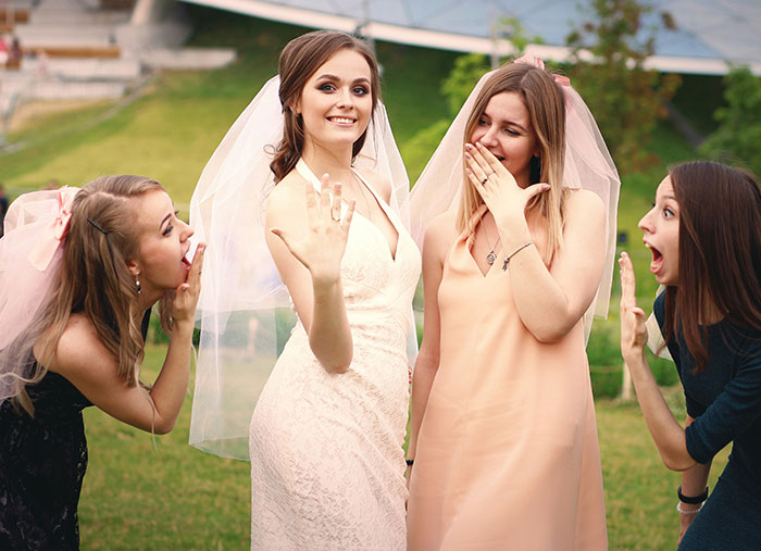Women in dresses outside, one in a wedding gown showing a ring to her surprised friends, theme of friendships ending.