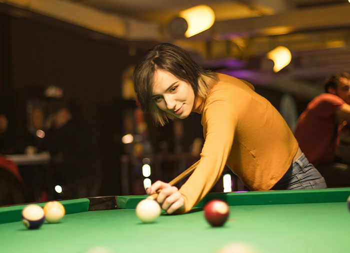 Woman in orange shirt playing pool, focused on a shot; social gathering environment, symbolizing great friendships.