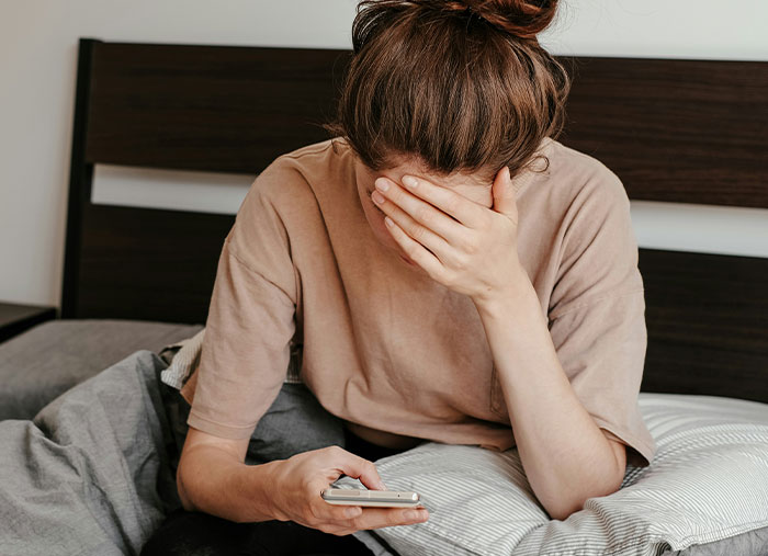 A person sitting on a bed, holding a phone and looking distressed about friendships.