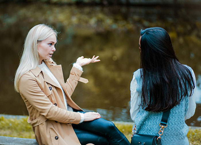 Two friends sitting by the water, one gesturing while in a conversation about ending friendships.